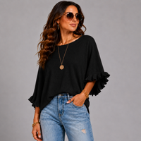 A woman modeling a black short-sleeve top with ruffled cuffs, paired with light blue jeans, gold jewelry, and sunglasses, against a solid gray studio background.