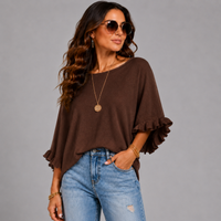 A woman wearing a brown short-sleeve top with ruffled cuffs and light blue jeans, posing against a solid gray studio background.